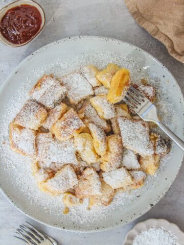 A forkful of torn pancake is held above into a pale blue ceramic plate of Austrian Kaiserschmarrn with jam on the side on a gray surface.