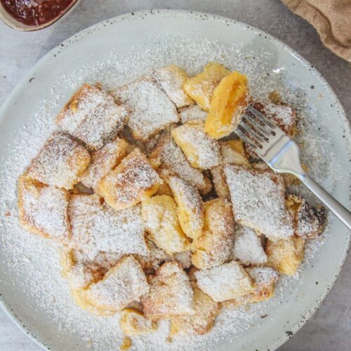 A forkful of torn pancake is held above into a pale blue ceramic plate of Austrian Kaiserschmarrn with jam on the side on a gray surface.