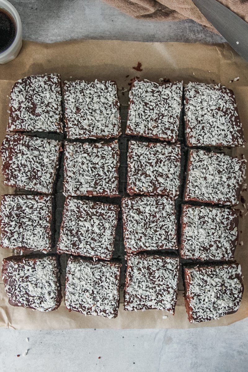 Swedish Karleksmums - Love Treats chocolate cake sits sliced up with frosting and desiccated coconut on top of parchment paper on a gray surface.