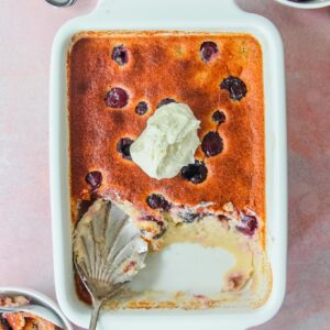 British Kentish Cherry Batter Pudding in a rectangle white baking dish with two individual servings on the sides on a light pink surface.