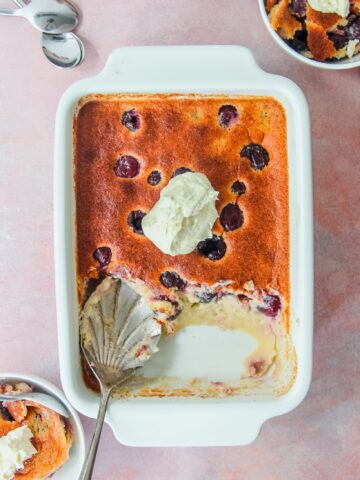 British Kentish Cherry Batter Pudding in a rectangle white baking dish with two individual servings on the sides on a light pink surface.