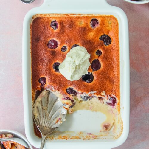 British Kentish Cherry Batter Pudding in a rectangle white baking dish with two individual servings on the sides on a light pink surface.
