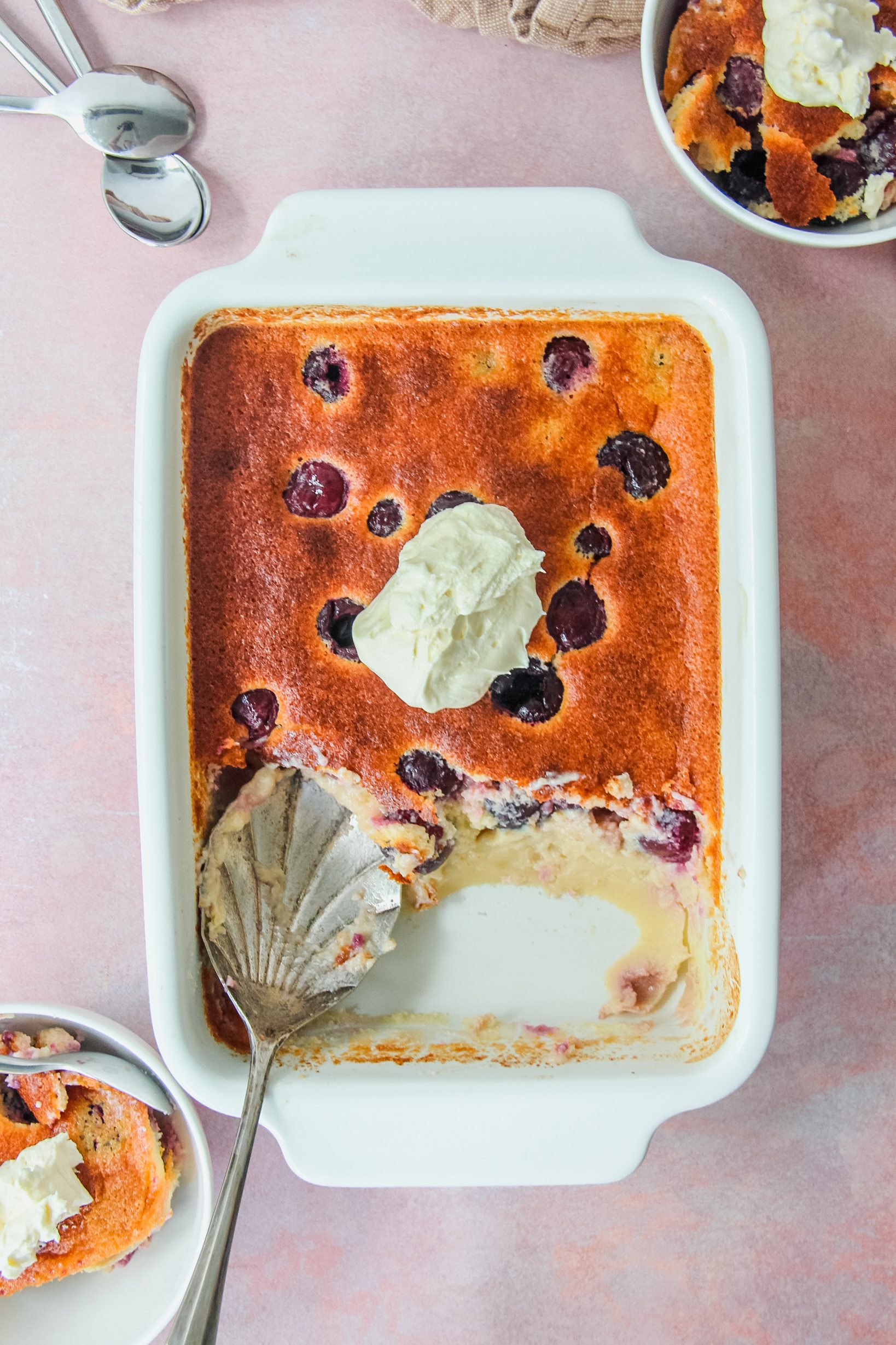 British Kentish Cherry Batter Pudding in a rectangle white baking dish with two individual servings on the sides on a light pink surface.