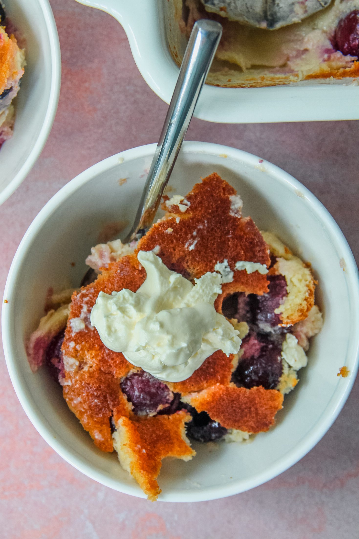 An individual serving of Kentish Cherry Batter Pudding sits in a white ceramic bowl on a light pink surface with a dollop of cream on top.