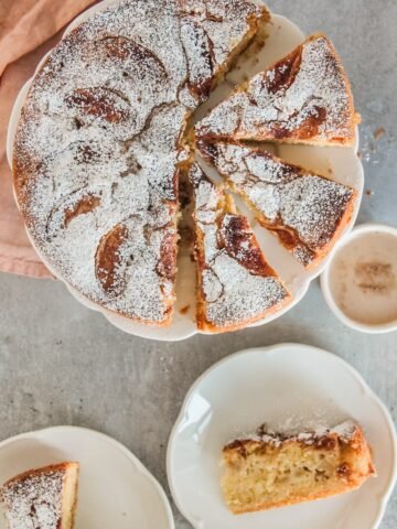 Kerry Apple Cake sits on a lightly scalloped apple cake with wedges of cake cut out and two pieces sitting on individual plates beneath the cake stand on a gray surface.