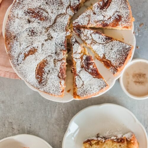 Kerry Apple Cake sits on a lightly scalloped apple cake with wedges of cake cut out and two pieces sitting on individual plates beneath the cake stand on a gray surface.