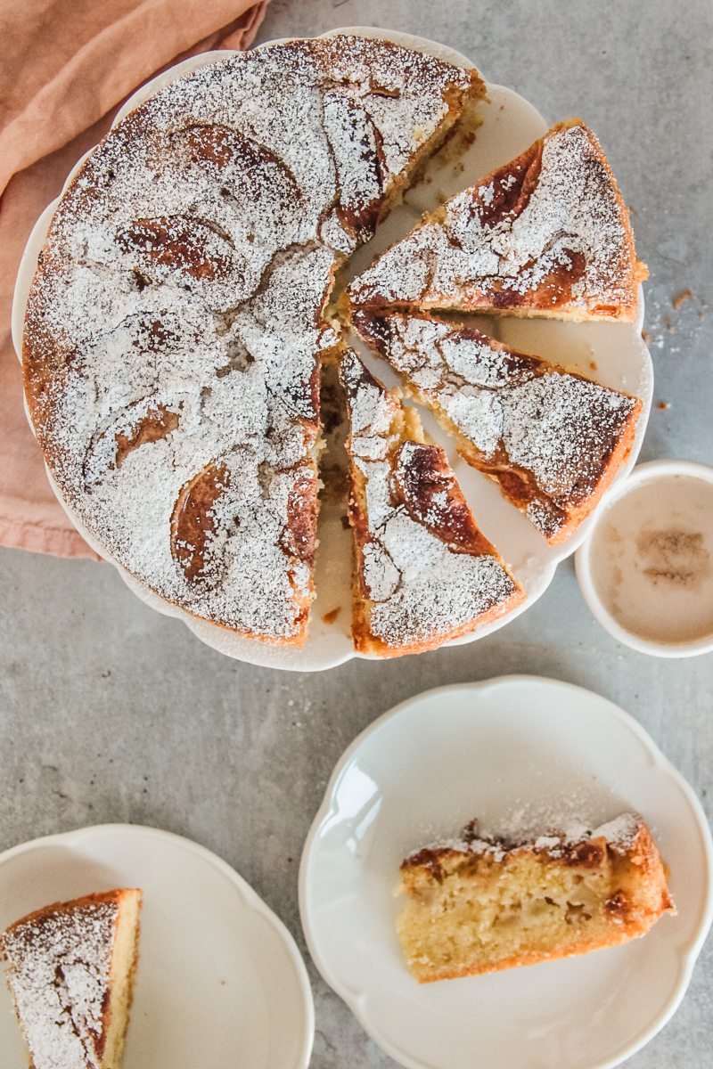 Kerry Apple Cake sits on a lightly scalloped apple cake with wedges of cake cut out and two pieces sitting on individual plates beneath the cake stand on a gray surface.