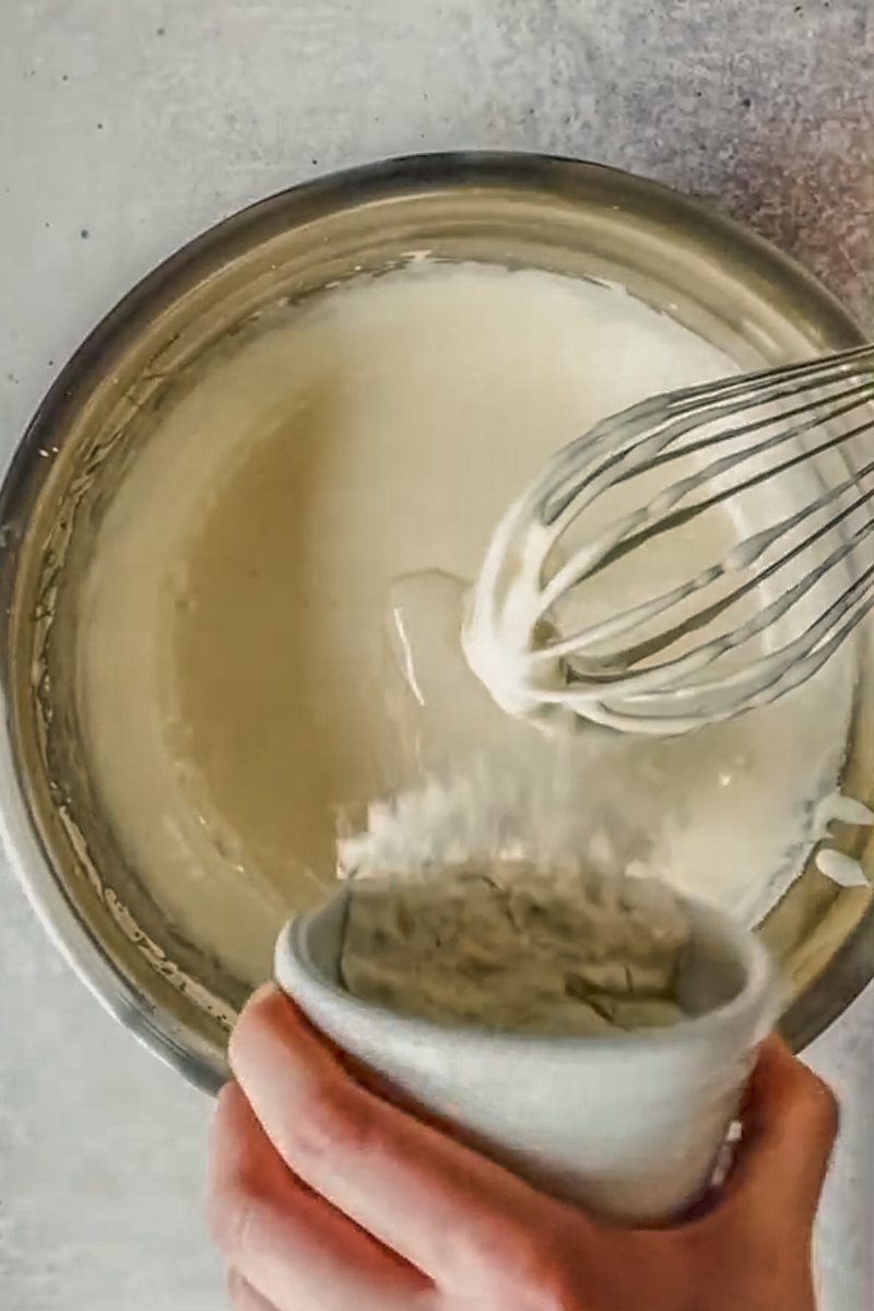 Flour is tossed into a whipped egg and sugar mixture in a stainless steel bowl on a gray surface.