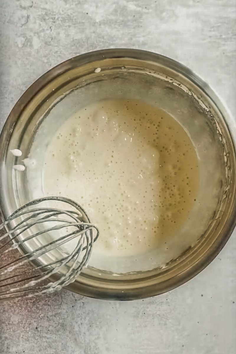 A pale sugar and egg mixture sits in a stainless steel bowl on a gray surface.