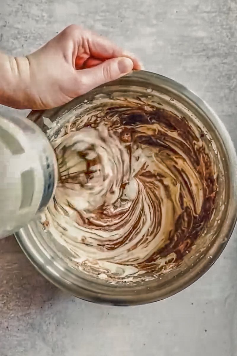 A cake batter is whisked in a stainless steel bowl on a gray surface.