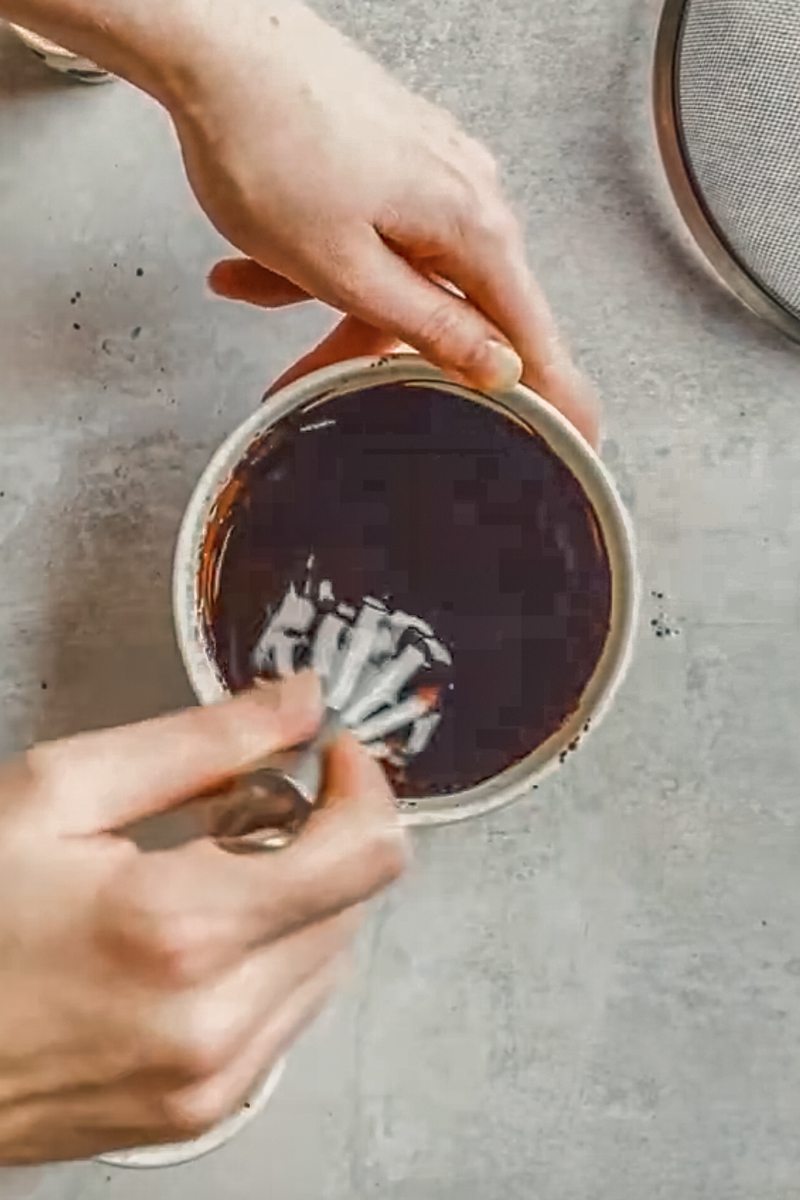 Melted butter and cocoa is wihisked in a white ceramic bowl on a gray surface.