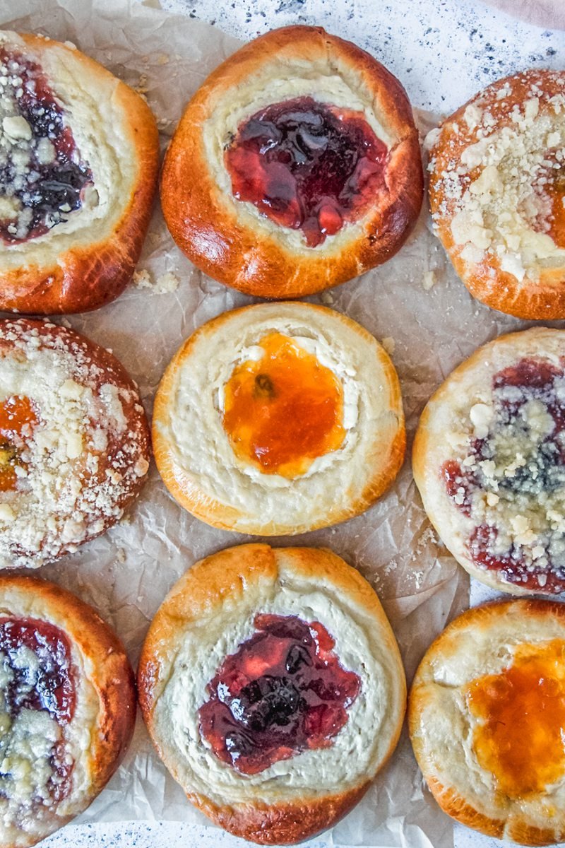 Kolaches with and without streusel toppings sit on parchment paper on a light gray surface.