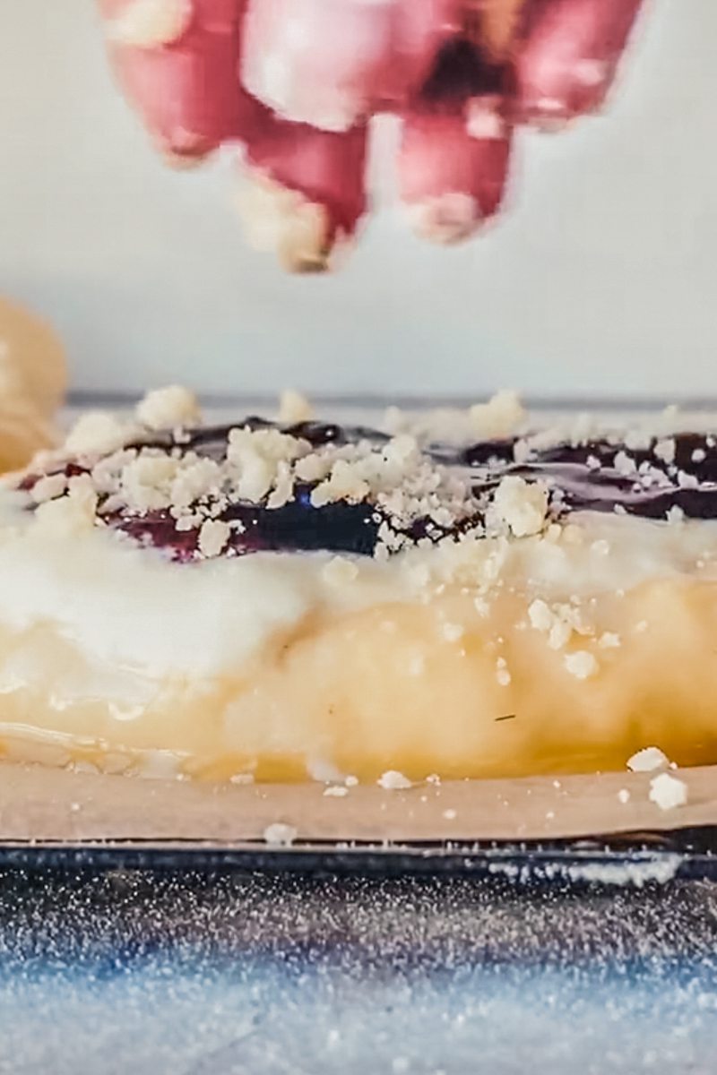 Streusel is sprinkled over kolache dough and fillings on a baking tray.