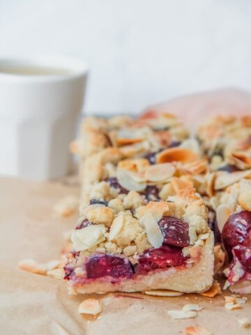 A close up of a slice of Latvian Cherry Crumb cake sits beside the remaining cake on parchment paper.