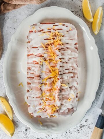 A lemon drizzle cake sits on a white scalloped plate on a gray surface.