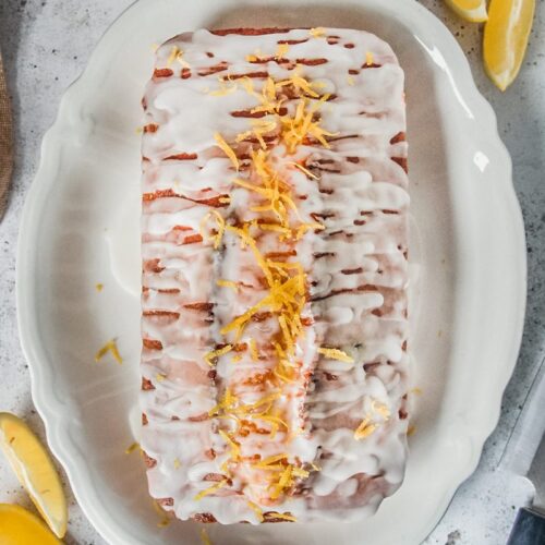 A lemon drizzle cake sits on a white scalloped plate on a gray surface.