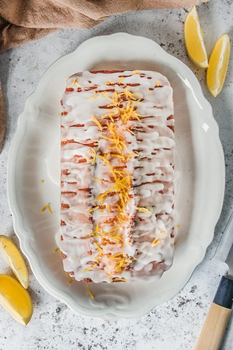 A lemon drizzle cake sits on a white scalloped plate on a gray surface.