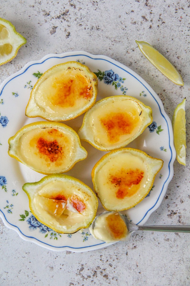 Lemon posset sits on a floral plate with caramelized tops on a light gray surface with lemon wedges beside.