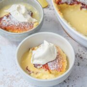 Lemon Surprise Pudding is served into individual ceramic bowls beside the remaining pudding in an oval white ceramic baking dish on a light gray surace.