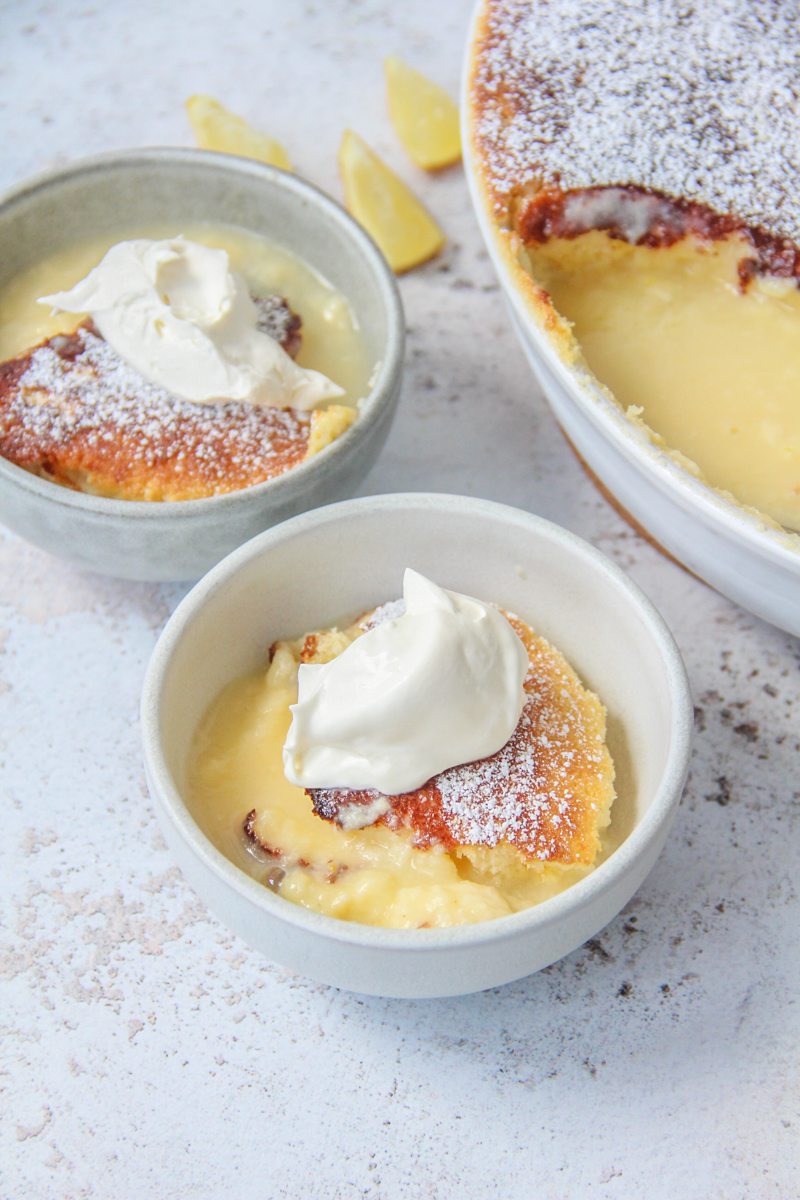 Lemon Surprise Pudding is served into individual ceramic bowls beside the remaining pudding in an oval white ceramic baking dish on a light gray surace.