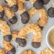 A close up of Mandelhoernchen - German Almond Horns lay on a light gray surface with coffee beside.