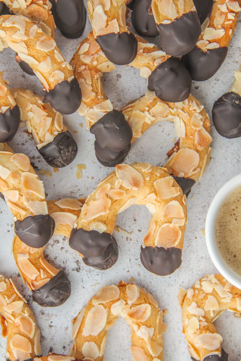 A close up of Mandelhoernchen - German Almond Horns lay on a light gray surface with coffee beside.