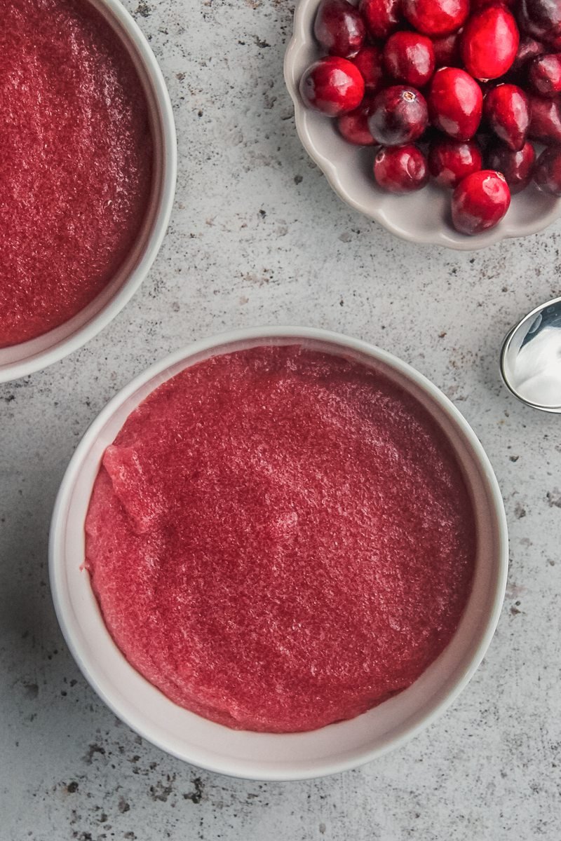 Mannavaht - Cranberry pudding sits in white ceramic bowls on a light gray surface with fresh cranberries sitting beside on a small plate.