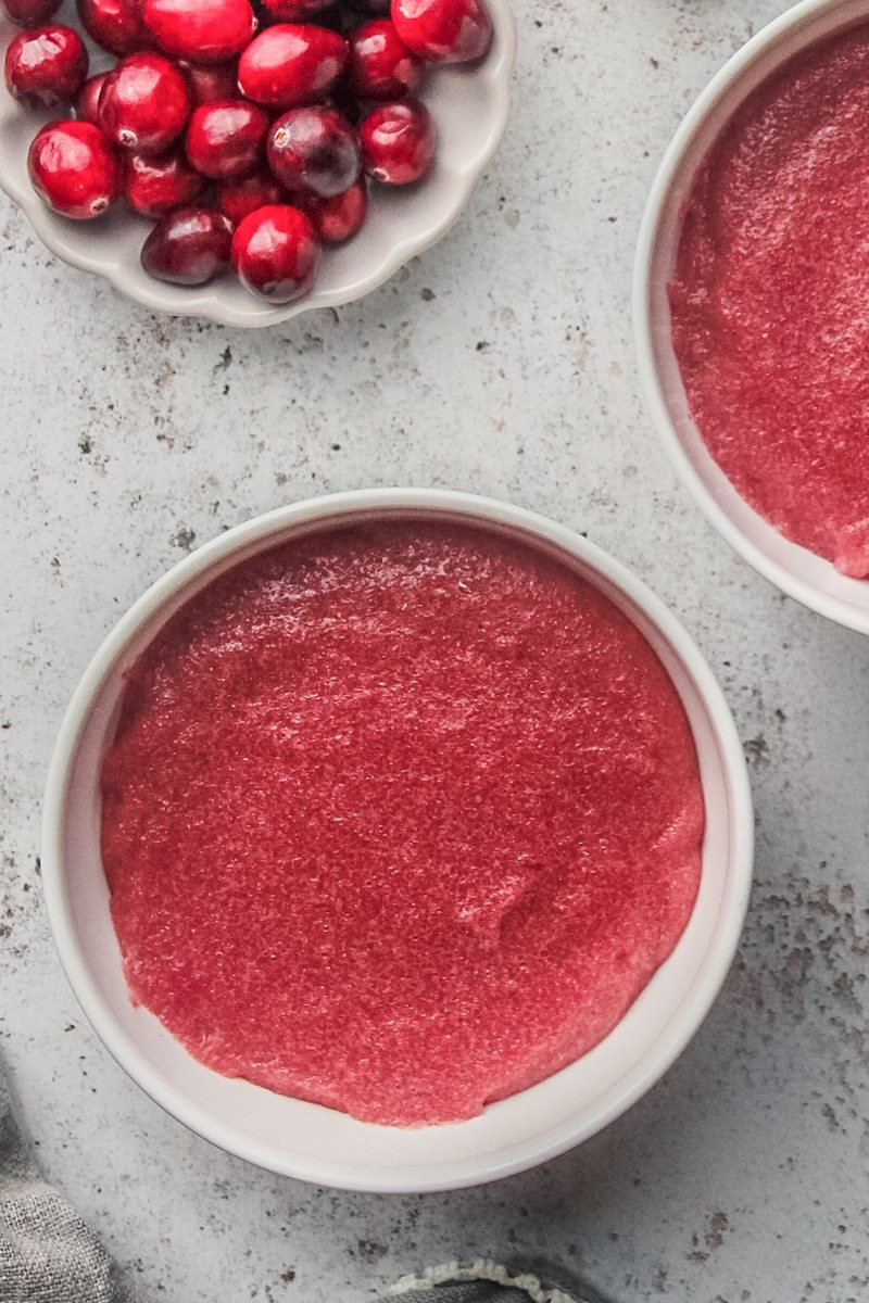 Estonian Mannavaht - cranberry pudding sits in white ceramic bowls on a light gray surface with fresh cranberries sitting beside on a small plate.