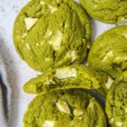 A matcha white chocolate cookie with a bite out of it sits between other matcha white chocolate cookies on a gray surface.