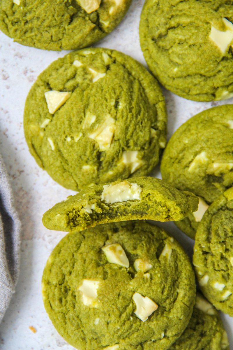 A matcha white chocolate cookie with a bite out of it sits between other matcha white chocolate cookies on a gray surface.