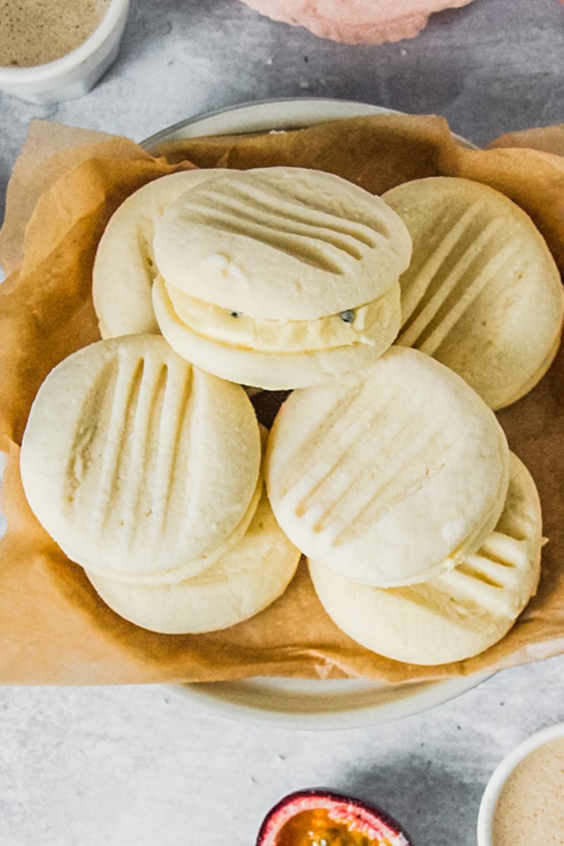 A stack of melting moments sandwich biscuits sit on a stack of rimmed ceramic plates on a gray surface.