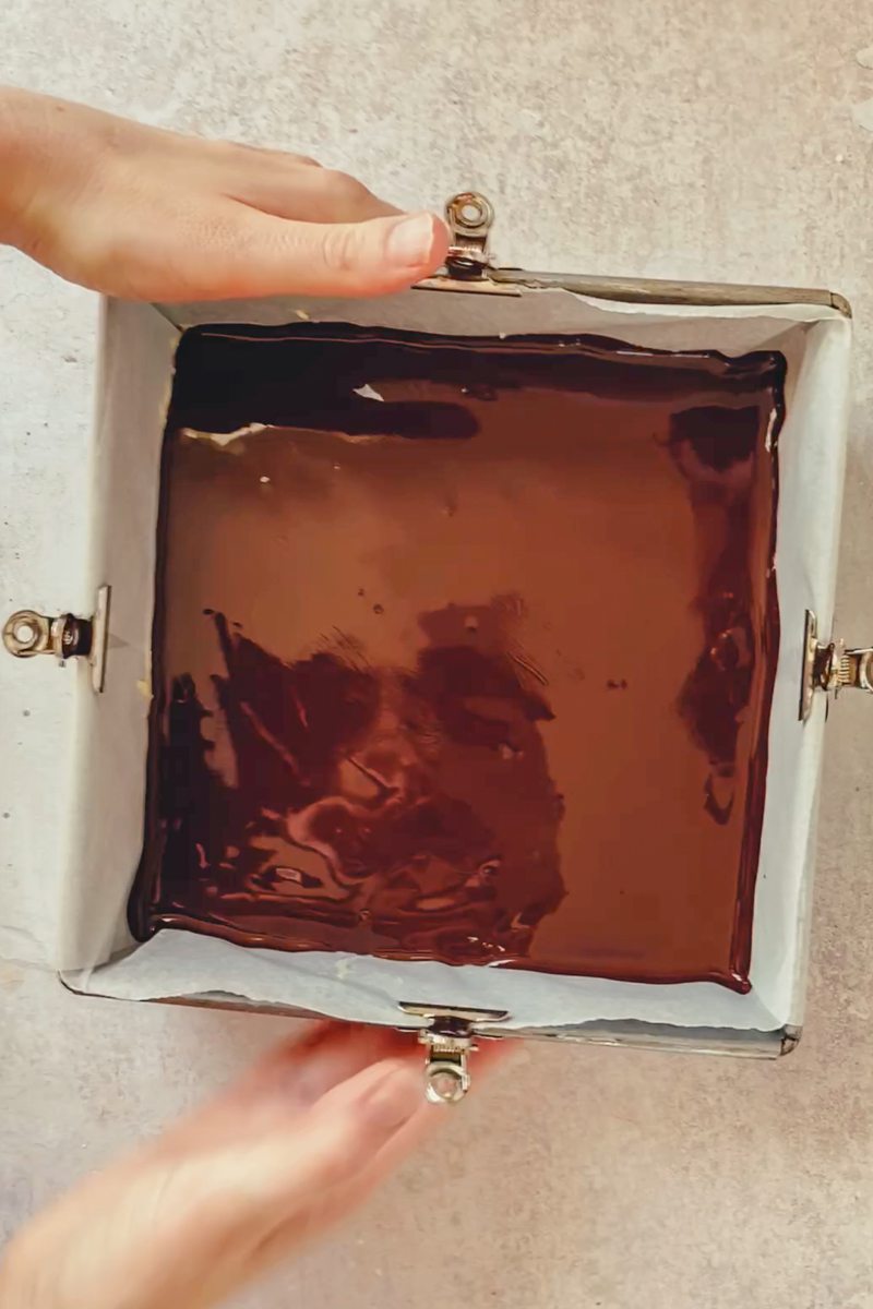 Glossy chocolate mixture on top of Nanaimo bars in a lined square metal baking tin held above a gray surface.