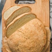 Native American masa bread sits on a wooden board with a few slices laying beside it on a gray cloth on a blue surface.