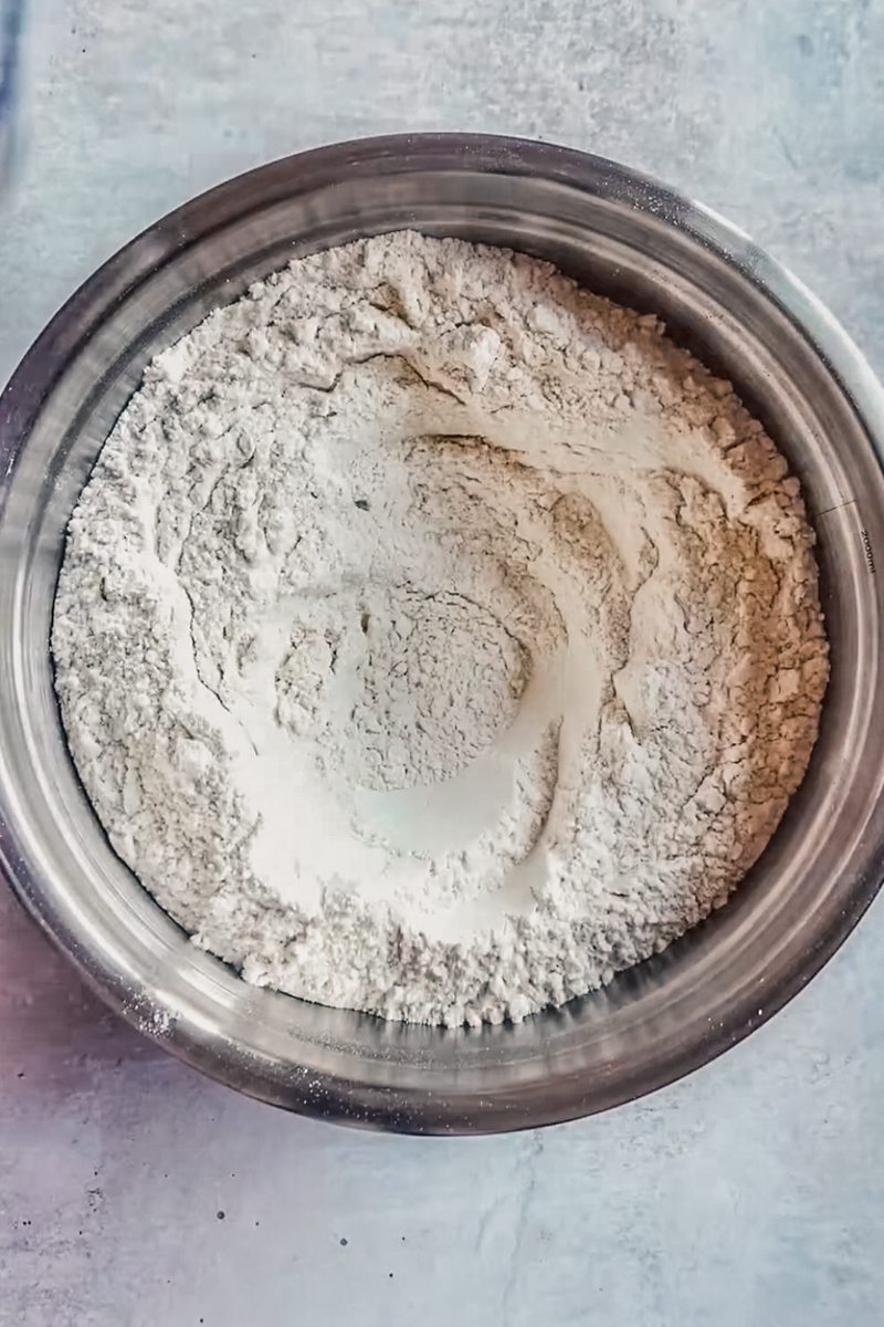 Dry ingredients sit in a stainless steel bowl with a well in the center on a gray surface.