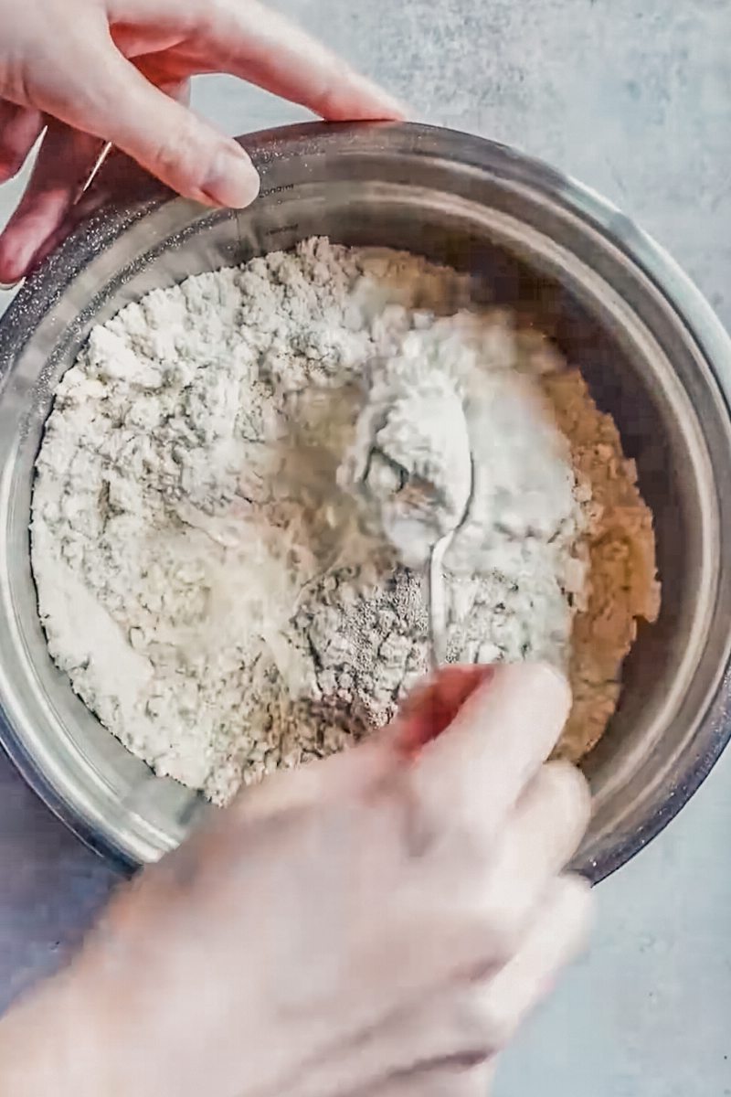 Dry ingredients and yeast are stirred in a stainless steel bowl on a gray surface.