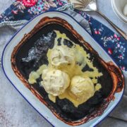 A baked chocolate pudding sits in a metal tin on a gray surface with scoops of melting vanilla ice cream on top.