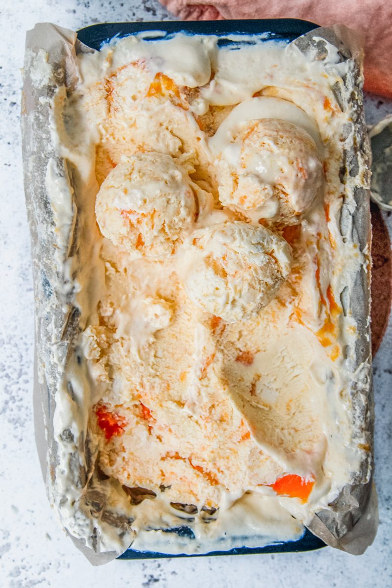 No Churn Peach Ice Cream sits scooped up in a lined rectangle metal loaf tin on a light gray surface.
