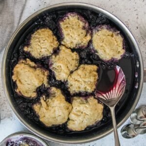 No bake blueberry cobbler is served up in the stainless steel pan its cooked in with a single serving sitting in a grey bowl on a light grey background.