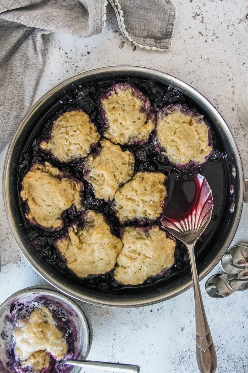 No bake blueberry cobbler is served up in the stainless steel pan its cooked in with a single serving sitting in a grey bowl on a light grey background.