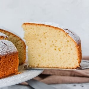 A slice of Spanish Olive Oil Cake sits on a cake serving knife with a brown cloth and the cake knife sitting on a light gray background beneath the cake stand.