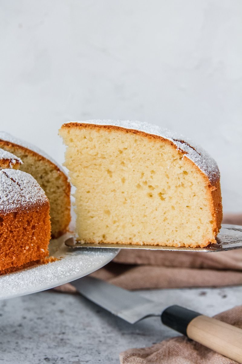 A slice of Spanish Olive Oil Cake sits on a cake serving knife with a brown cloth and the cake knife sitting on a light gray background beneath the cake stand.