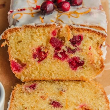 A slice of orange cranberry cake leans up against the loaf with cranberries throughout the cake batter on a wooden board.