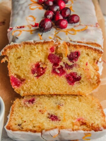 A slice of orange cranberry cake leans up against the loaf with cranberries throughout the cake batter on a wooden board.