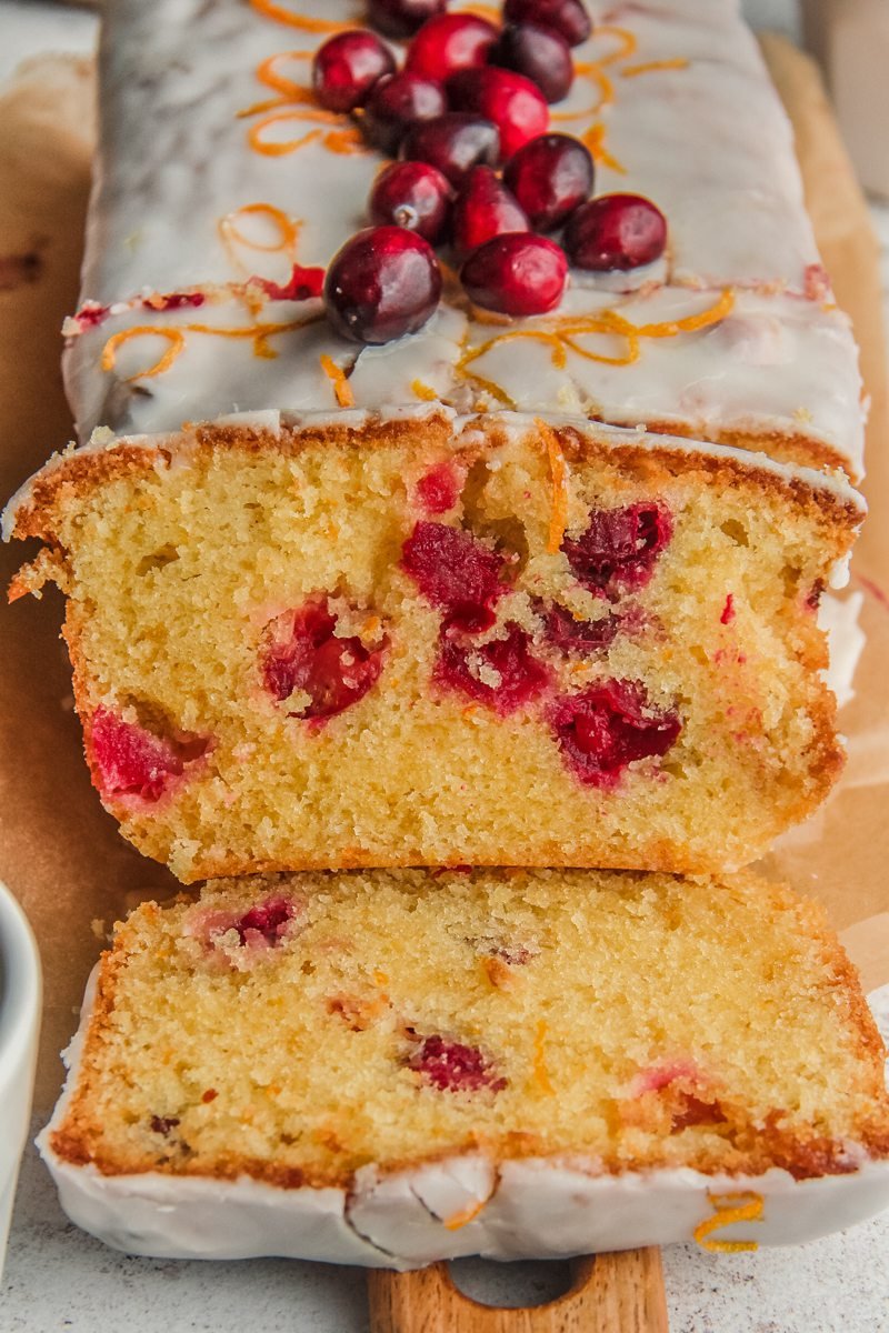 A slice of orange cranberry cake leans up against the loaf with cranberries throughout the cake batter on a wooden board.