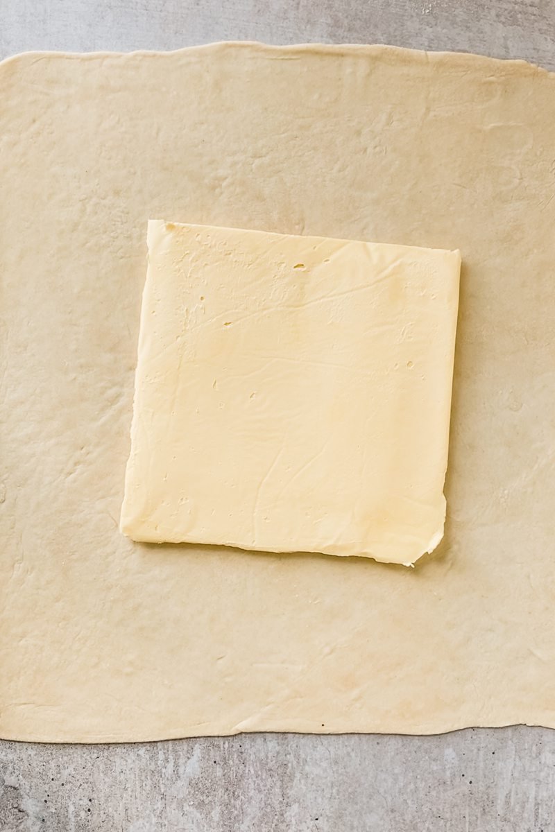 A square piece of butter sits in the center of pastry dough on a gray surface.