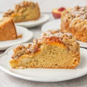 A slice of Peach Crumb Cake sits on a round white ceramic plate on a gray surface with remaining slices behind.