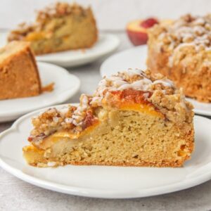 A slice of Peach Crumb Cake sits on a round white ceramic plate on a gray surface with remaining slices behind.