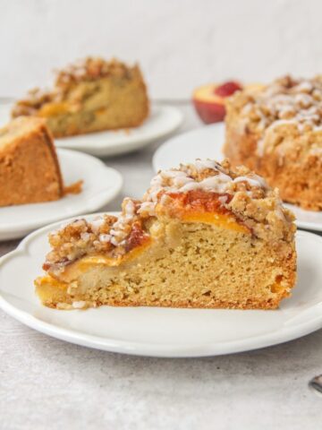 A slice of Peach Crumb Cake sits on a round white ceramic plate on a gray surface with remaining slices behind.