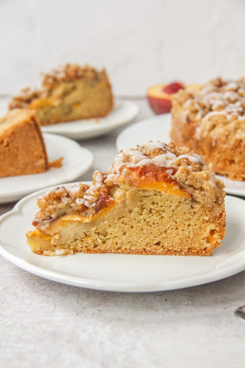 A slice of Peach Crumb Cake sits on a round white ceramic plate on a gray surface with remaining slices behind.