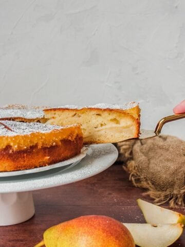 A slice of French pear cake is lifted from the cake plate sitting on a cake stand on a wooden background with a brown cloth in the background and pear in the foreground.